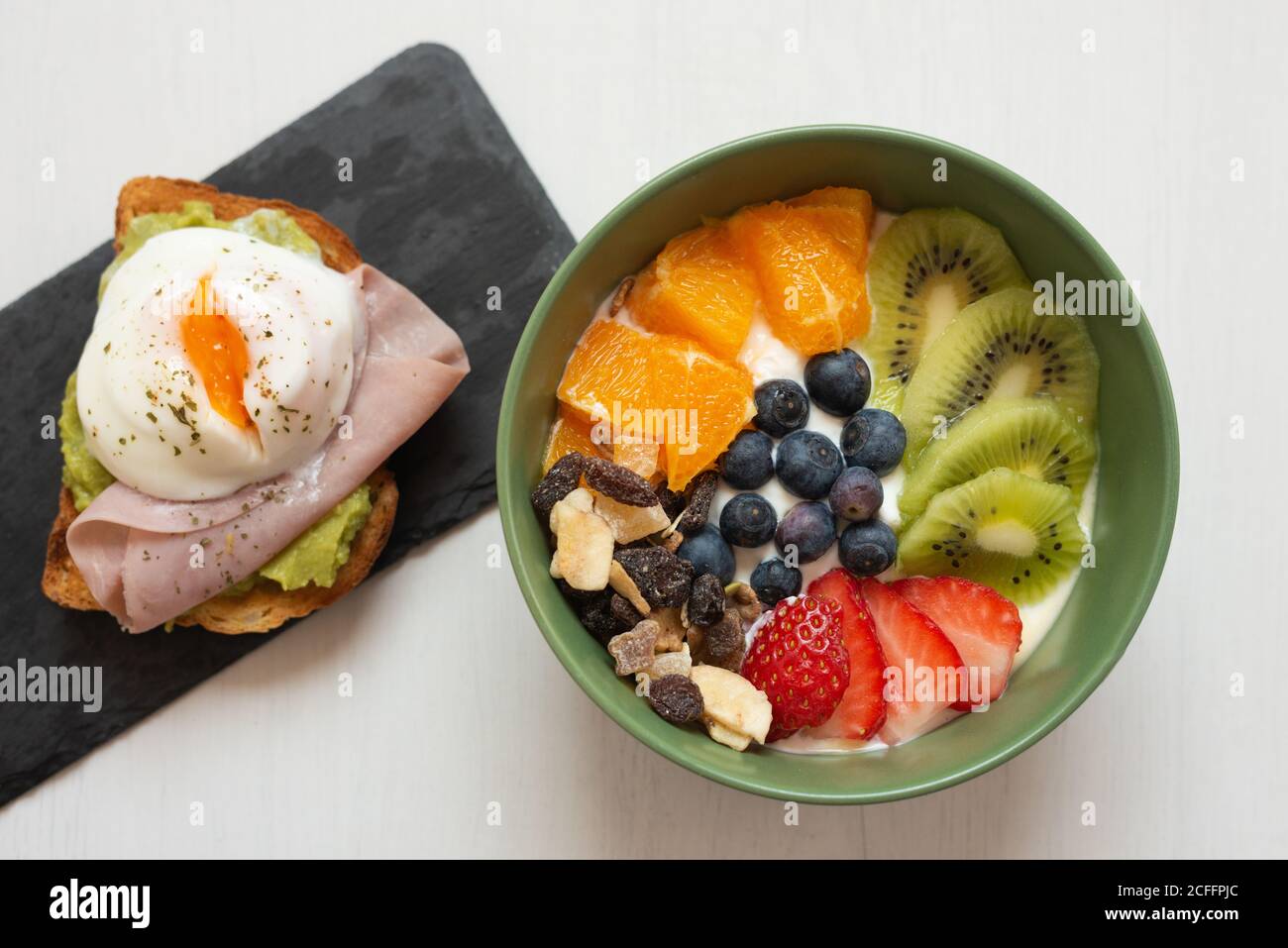 Dall'alto della gustosa ciotola per la colazione con frutta a fette e. bacche poste sul tavolo vicino al pane tostato con uovo fritto e. prosciutto Foto Stock