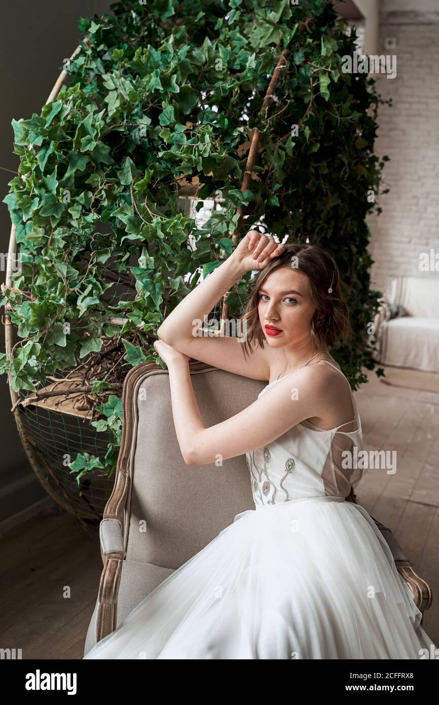 Donna splendida con labbra rosse in abito bianco seduta su poltrona Foto Stock