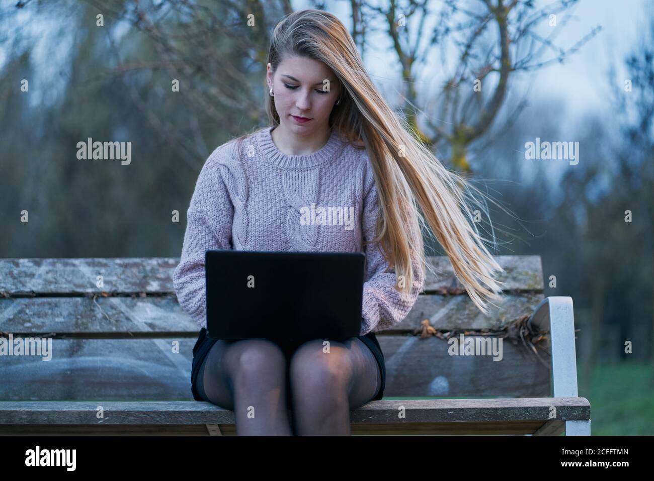 Giovane donna in abbigliamento casual e con capelli che scorre seduti sulla panca nel parco e rispondendo alle e-mail mentre si digita sulla tastiera del computer portatile Foto Stock