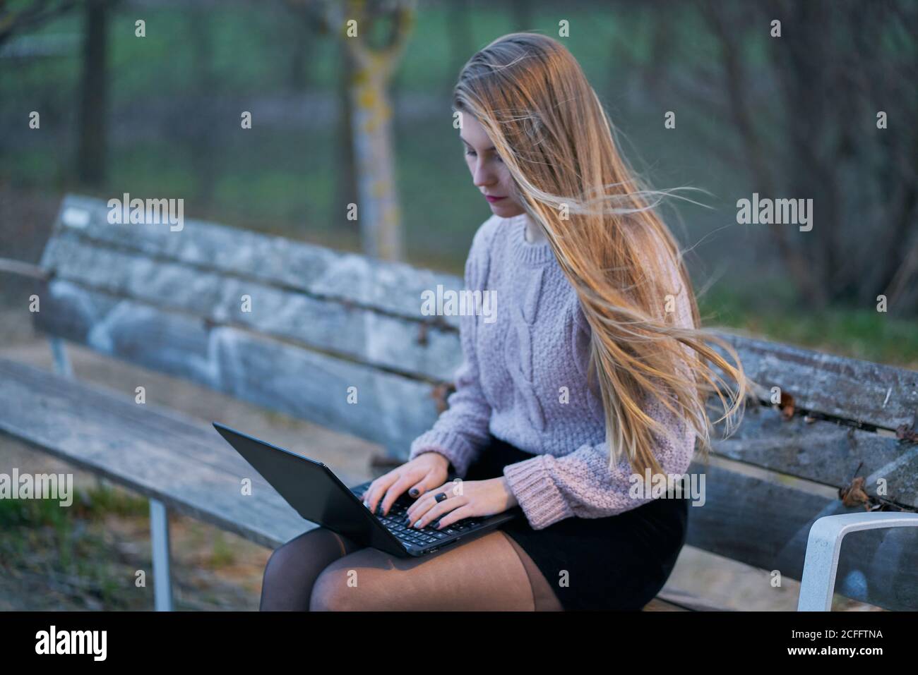Giovane donna in abbigliamento casual e con capelli che scorre seduti sulla panca nel parco e rispondendo alle e-mail mentre si digita sulla tastiera del computer portatile Foto Stock
