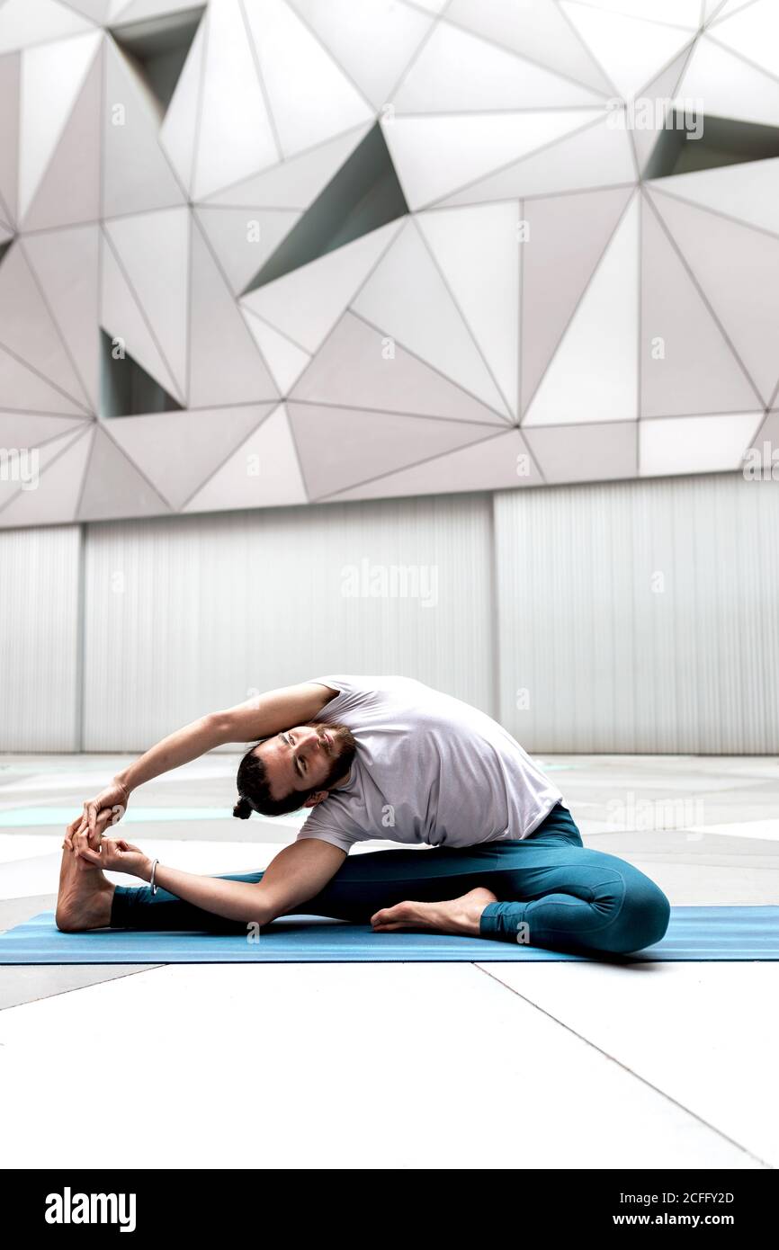 Barefoot sopportato ragazzo facendo Revoluto testa a ginocchio esercizio mentre seduta sul tappetino contro la parete geometrica durante l'allenamento yoga Foto Stock
