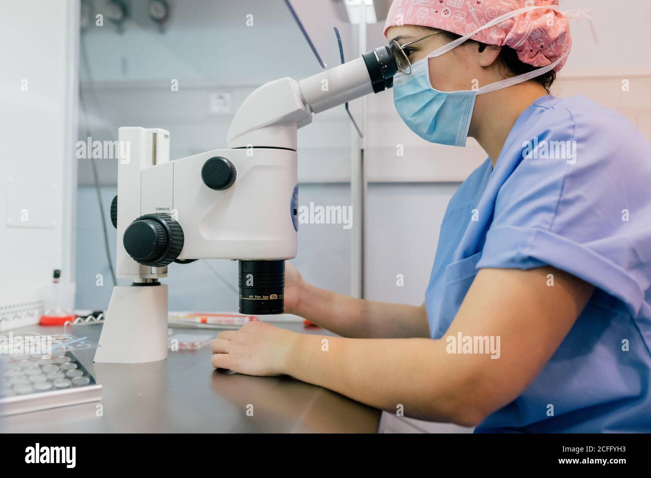 Vista laterale della donna in uniforme medica e maschera utilizzando un microscopio moderno per esaminare le cellule umane mentre si lavora in laboratorio di clinica moderna Foto Stock