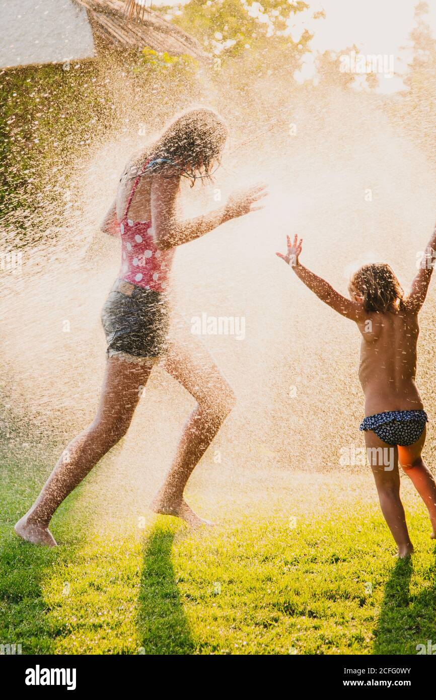 Adolescente anonimo a corpo pieno e bambina che corre e gioca in gocce di spruzzatura acqua pulita mentre ci si diverte giardino insieme Foto Stock