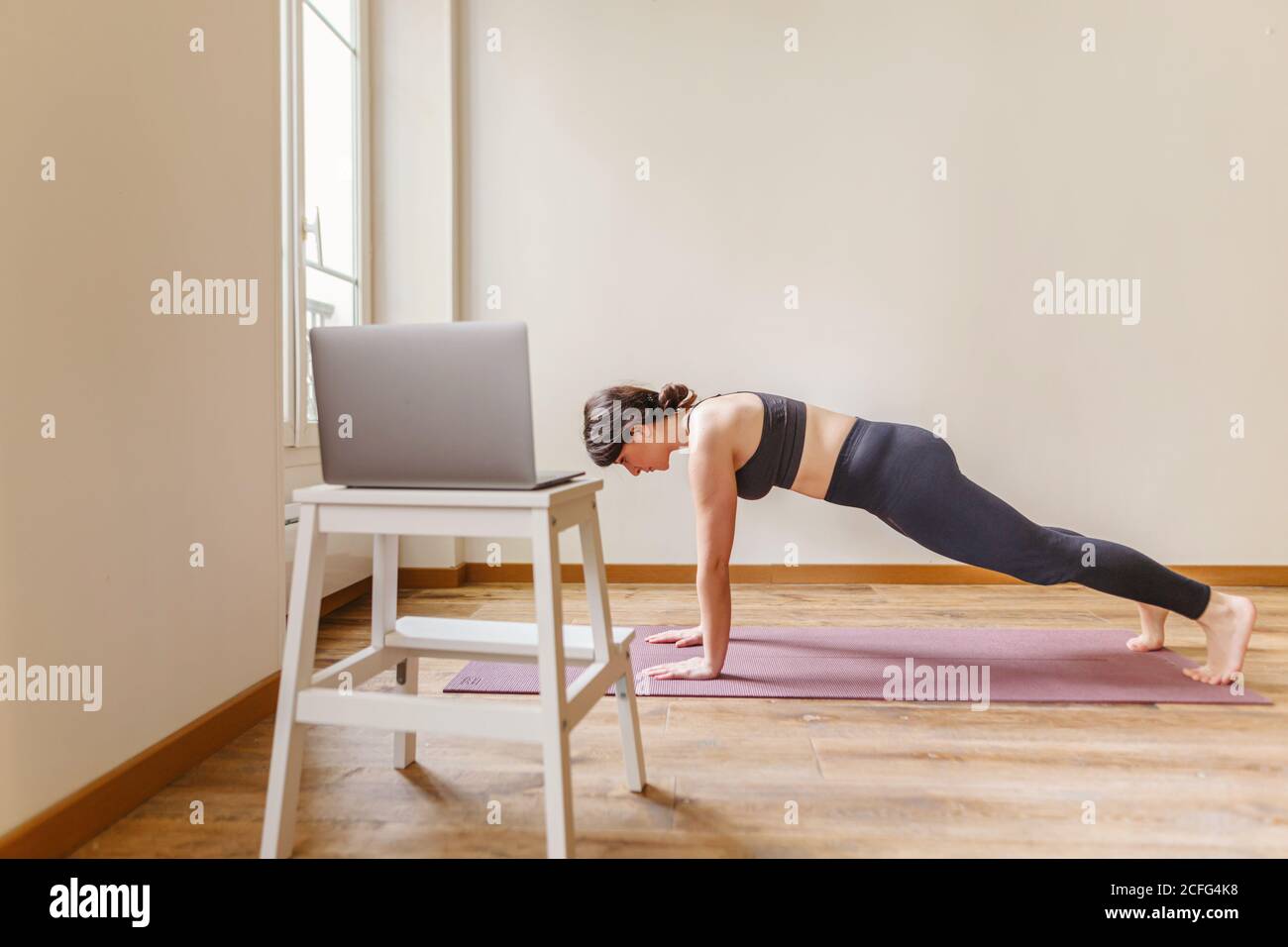 Vista laterale di Donna in vestito sportivo in piedi in tavola posa sul tappetino vicino a sgabello con computer portatile in appartamento il giorno di sole Foto Stock