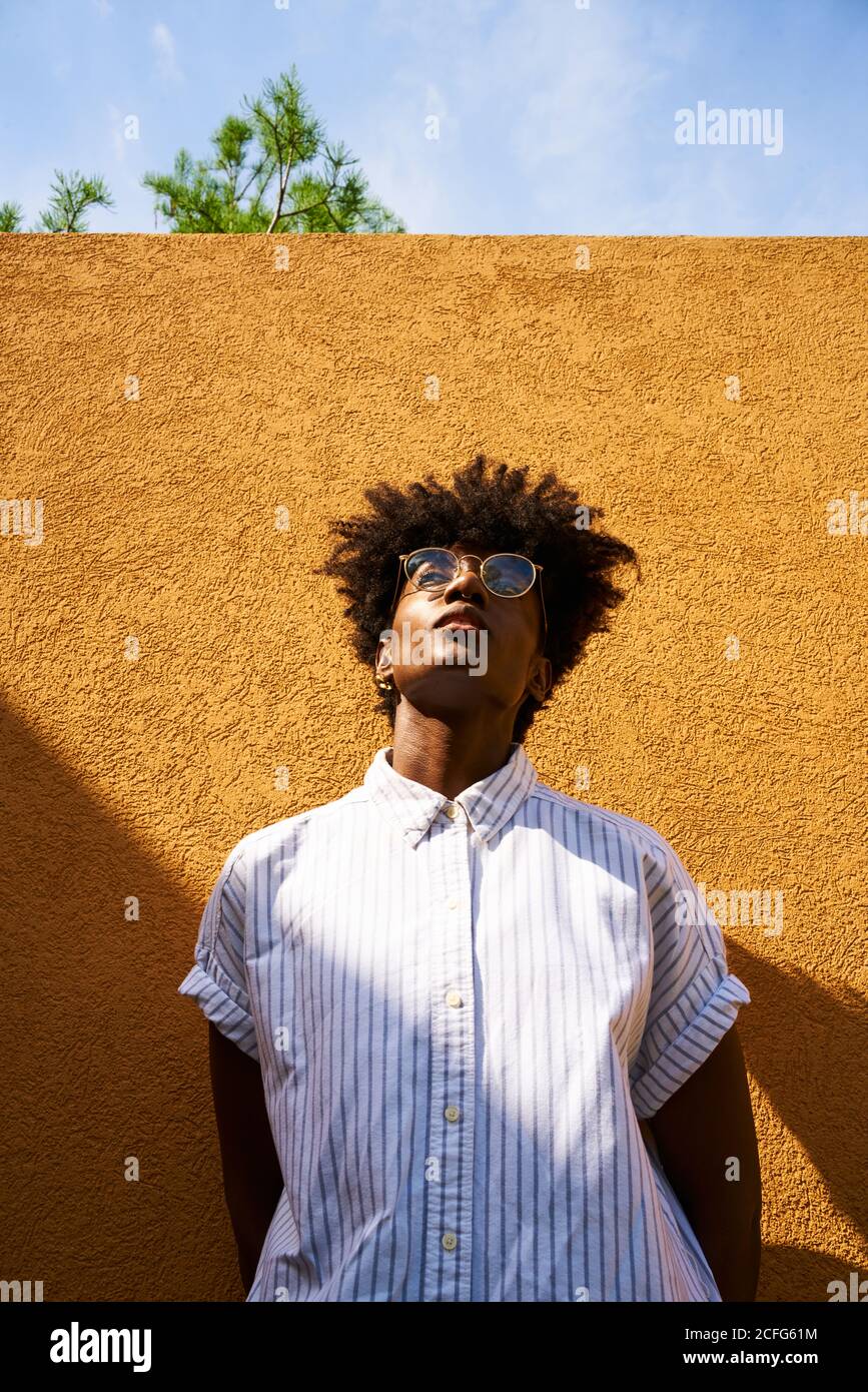 Tranquilla femmina nera in occhiali rotondi e camicia bianca verso l'alto mentre si sta sulla strada in travi a vista contro intonacato giallo parete di un edificio moderno Foto Stock