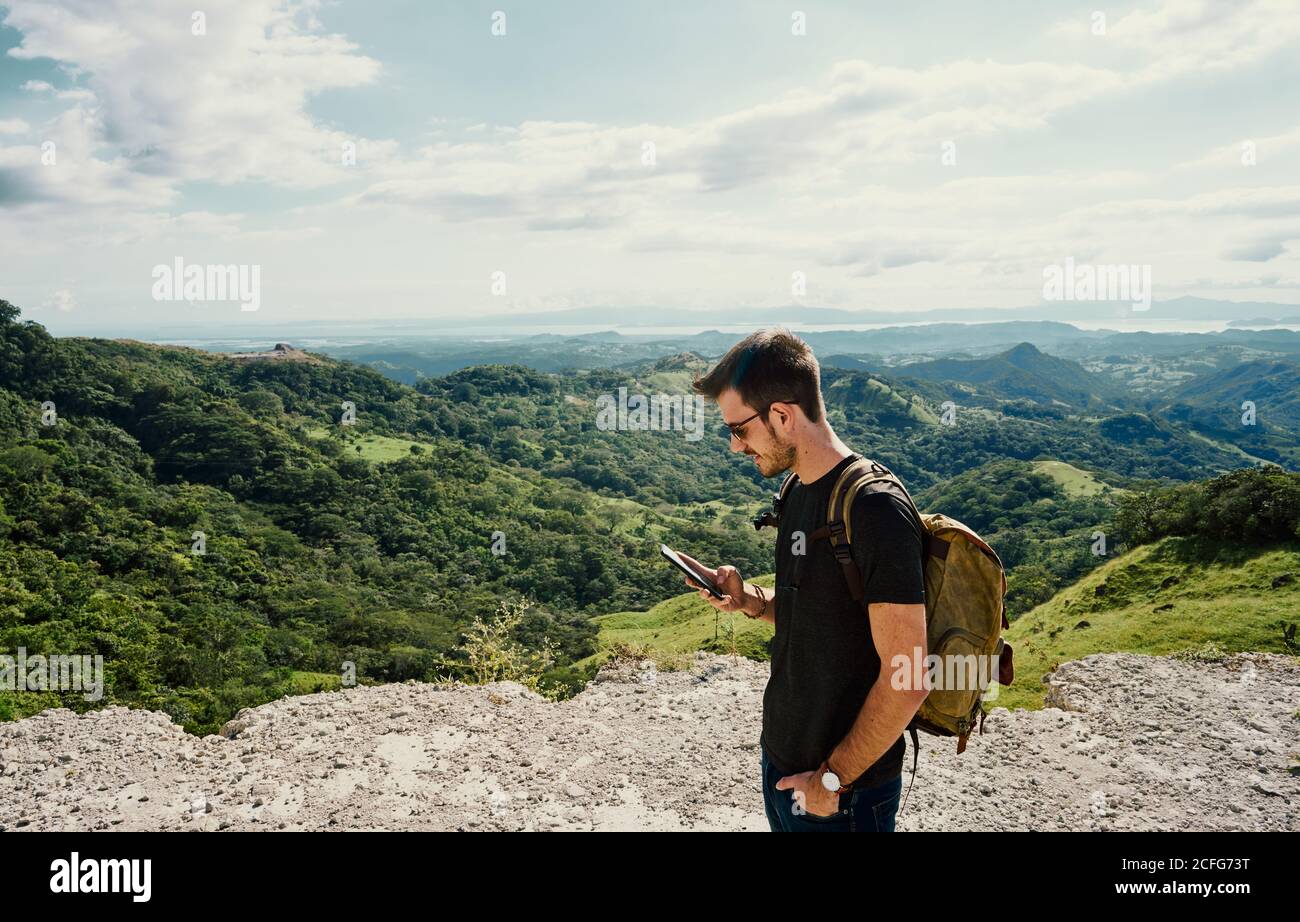 Vista laterale dell'escursionista maschile concentrato con messaggi di testo zaino Sul cellulare durante il trekking in località di montagna in Costa Rica Foto Stock