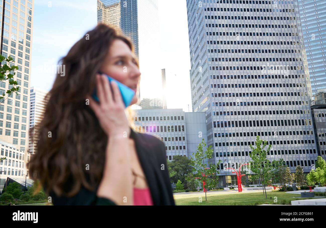 Donna d'affari soddisfatta in giacca elegante che discute le edizioni di lavoro mentre parla sul telefono cellulare ed in piedi sullo sfondo sfocato dei grattacieli Foto Stock