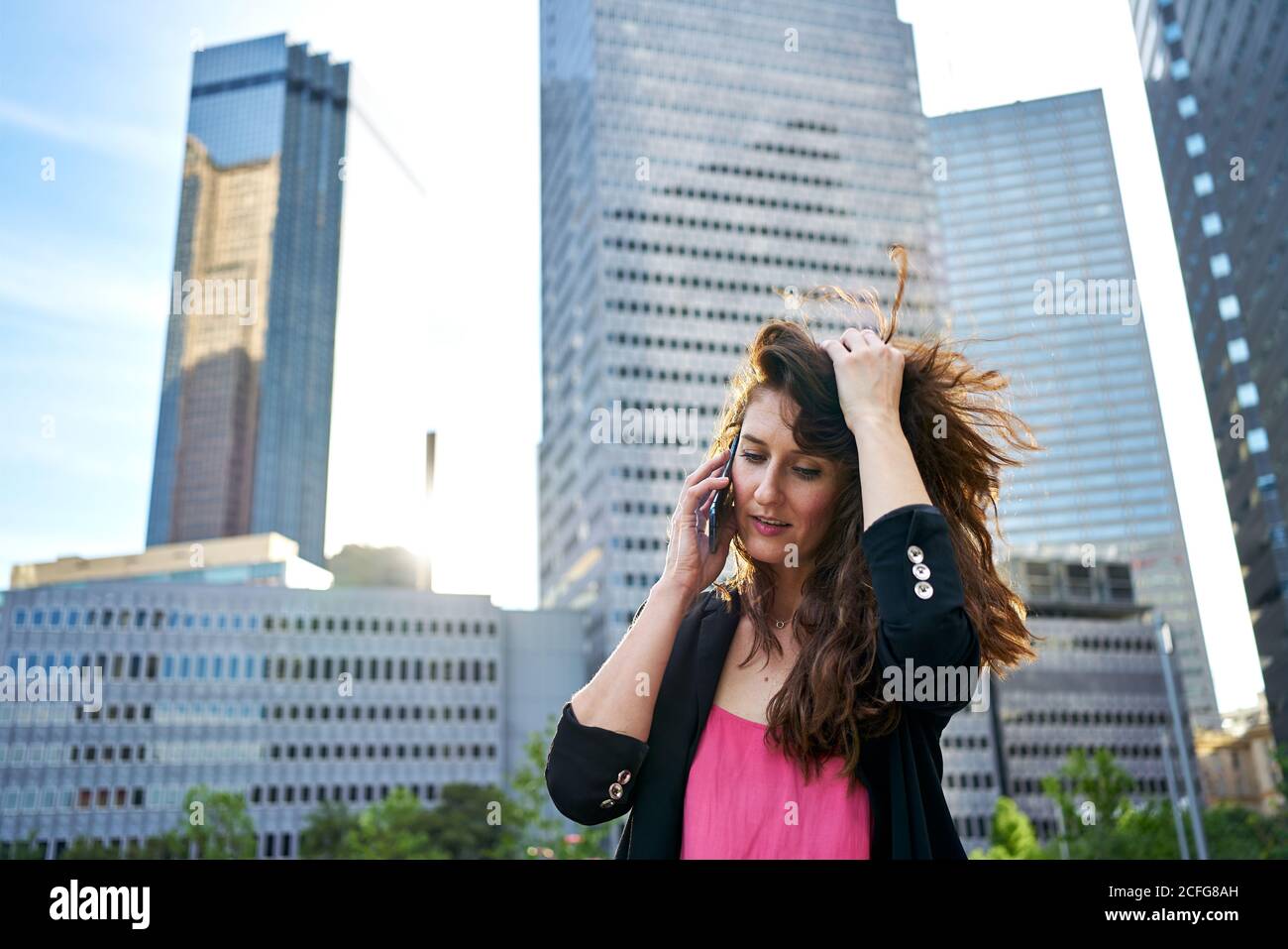 Donna d'affari soddisfatta in giacca elegante che discute le edizioni di lavoro mentre parla sul telefono cellulare ed in piedi sullo sfondo sfocato dei grattacieli Foto Stock