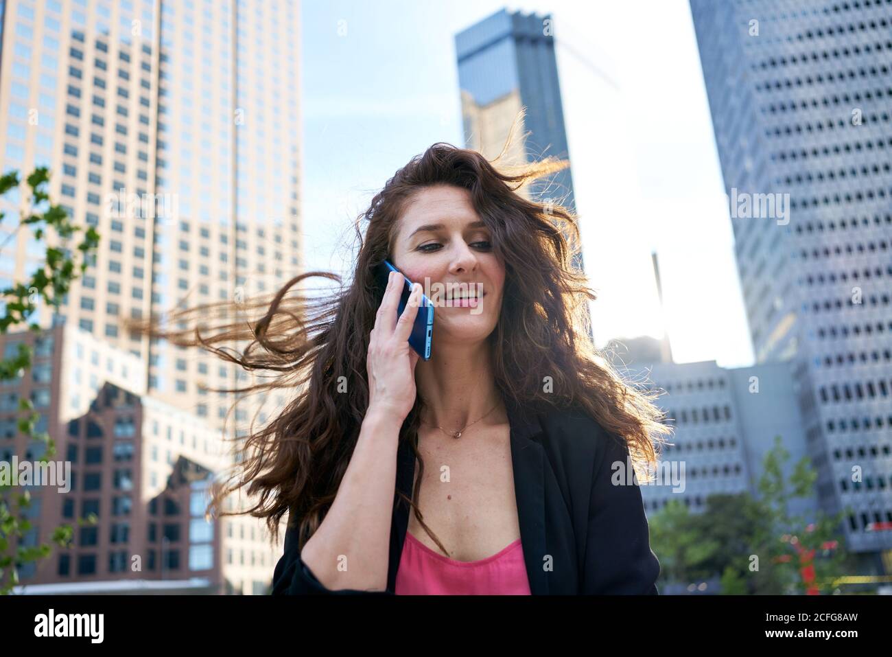 Donna d'affari soddisfatta in giacca elegante che discute le edizioni di lavoro mentre parla sul telefono cellulare ed in piedi sullo sfondo sfocato dei grattacieli Foto Stock