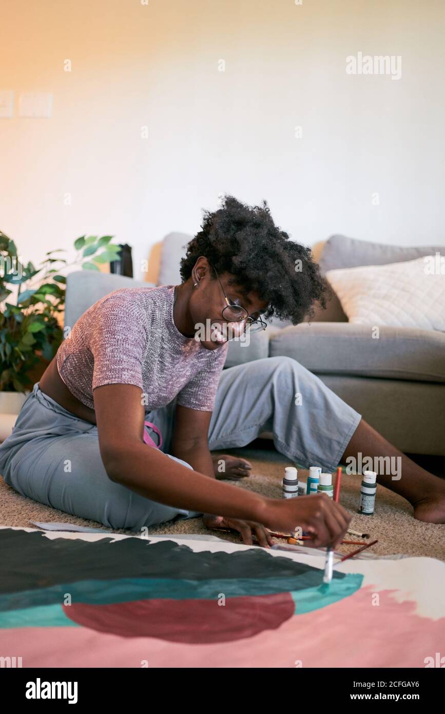 Giovane bella donna afroamericana con capelli afro, artista amatoriale in abito casual disegno colorato immagine con pennello su carta mentre seduto sul pavimento in accogliente appartamento Foto Stock