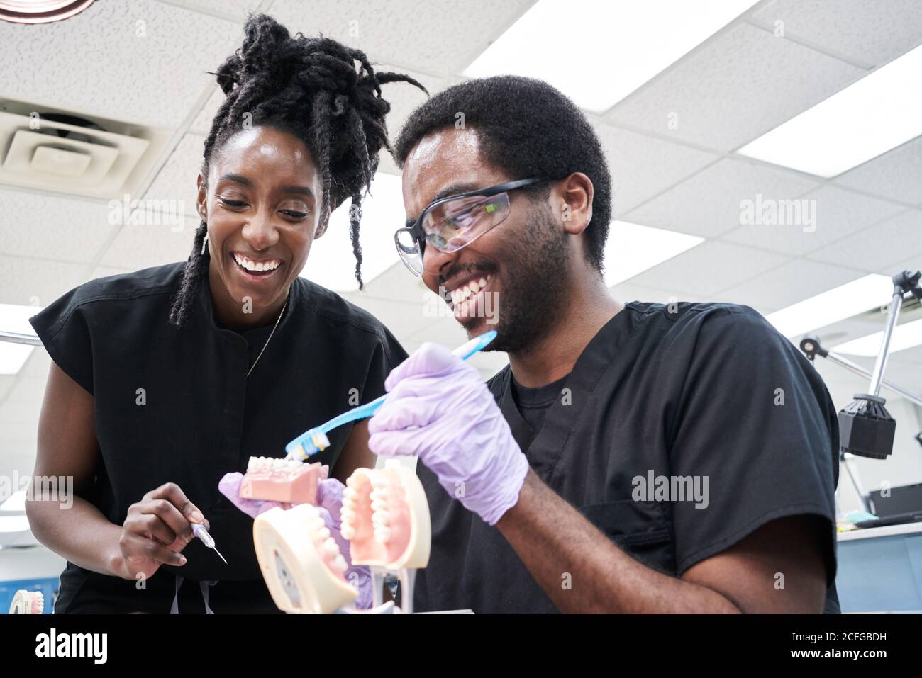 Angolo basso di felice Afro-American Woman con greggi e barbuto uomo sorridendo e spazzolando falsi denti durante il lavoro in laboratorio Foto Stock
