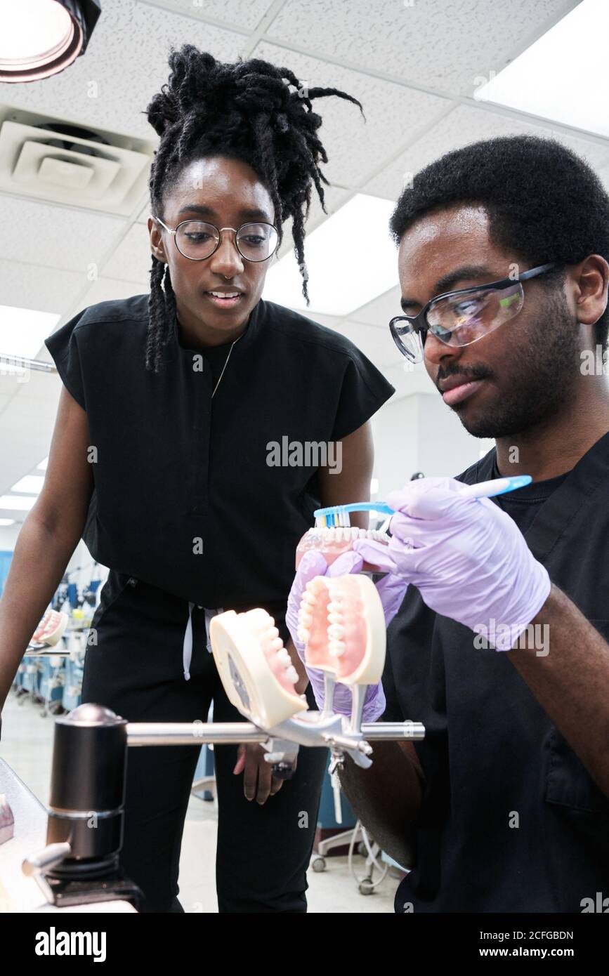 Angolo basso di felice Afro-American Woman con greggi e barbuto uomo sorridendo e spazzolando falsi denti durante il lavoro in laboratorio Foto Stock