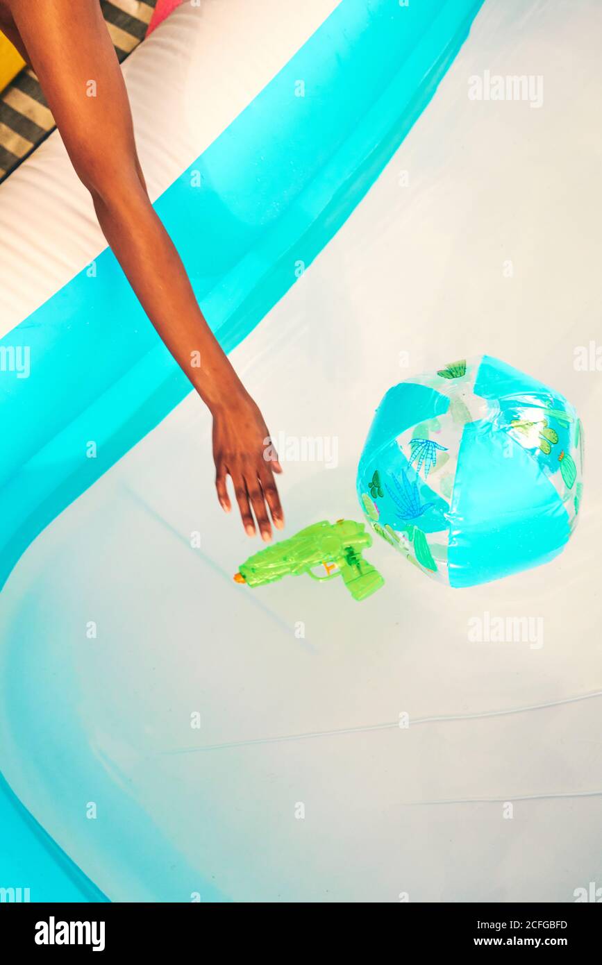 Dall'alto di femmina nera che tocca l'acqua in piscina con pistola ad acqua in plastica e palla da spiaggia Foto Stock