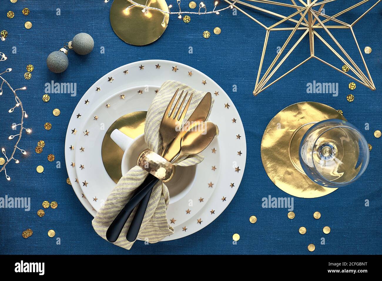 Tavolo di Natale con piatti bianchi, utensili dorati e decorazioni natalizie. Vista dall'alto su sfondo in tessuto di lino blu scuro. Luci di Natale garl Foto Stock