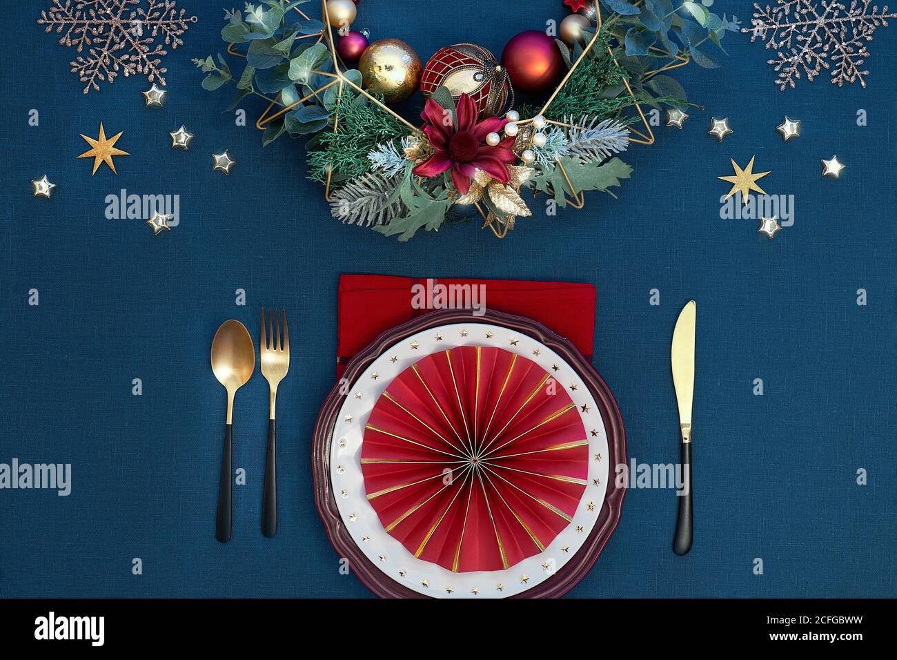 Tavolo natalizio con piatti bianchi, utensili dorati e decorazioni dorate di colore rosso scuro. Vista dall'alto su classico fondo tessile di lino blu. Foto Stock
