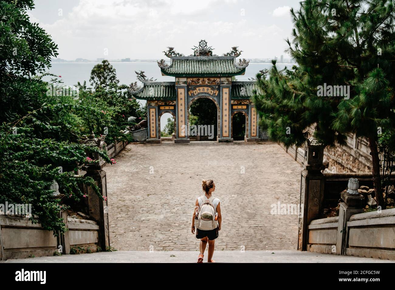 Vista posteriore di turista femminile anonimo con zaino in piedi scale di fronte all'arco del tempio buddista in Vietnam Foto Stock