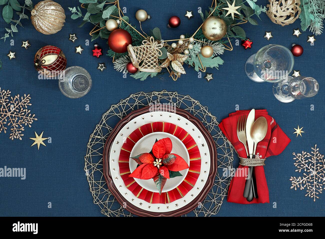 Tavolo natalizio con piatti bianchi, utensili dorati e decorazioni natalizie dorate di colore rosso scuro. Disposizione piatta, vista dall'alto su tovaglia di tessuto di lino blu. Foto Stock