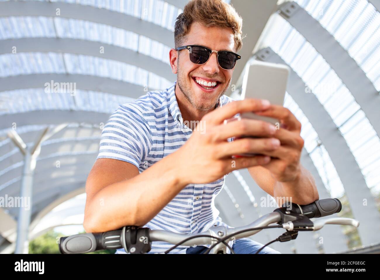 Da sotto colpo di giovane ragazzo attraente in occhiali da sole che si inclinano sul manubrio della bicicletta e smartphone di navigazione in un percorso coperto Foto Stock