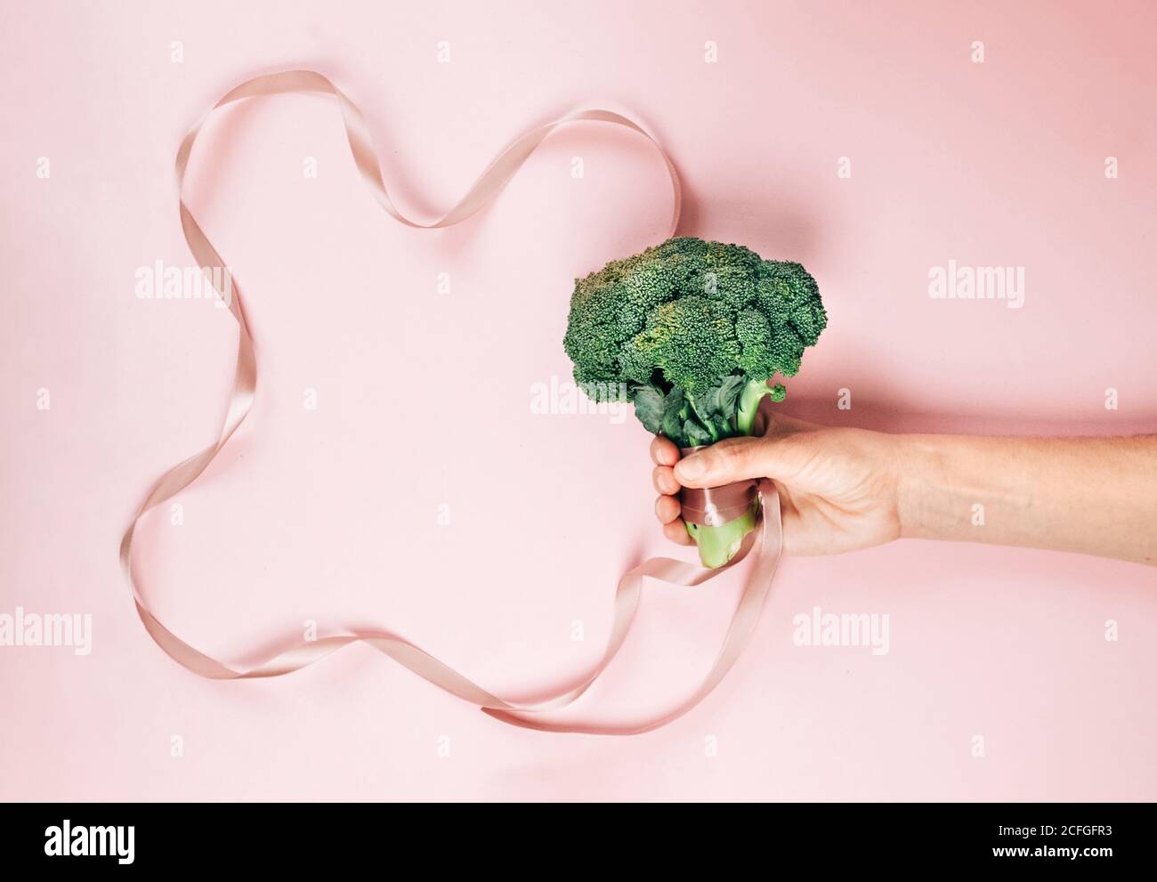 Vista dall'alto di una persona irriconoscibile che mostra broccoli maturi con seta nastro come simbolo di dieta e stile di vita sano contro il rosa sfondo Foto Stock