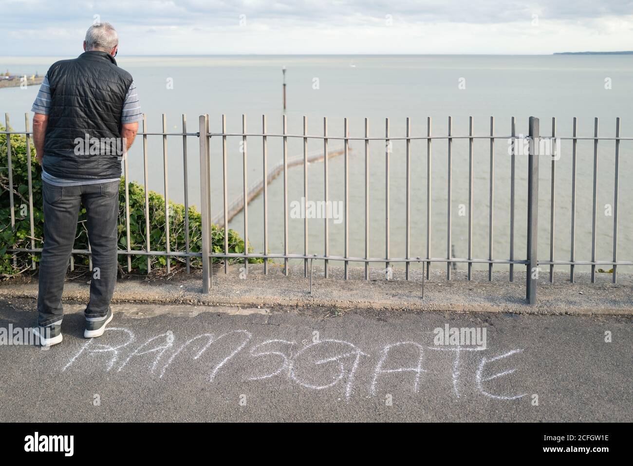 Un uomo sorge su una passeggiata in cima a una scogliera che si affaccia su una recinzione verso il mare sottostante. Il nome della città Ramsgate è stato calato sulla asfalto. Foto Stock