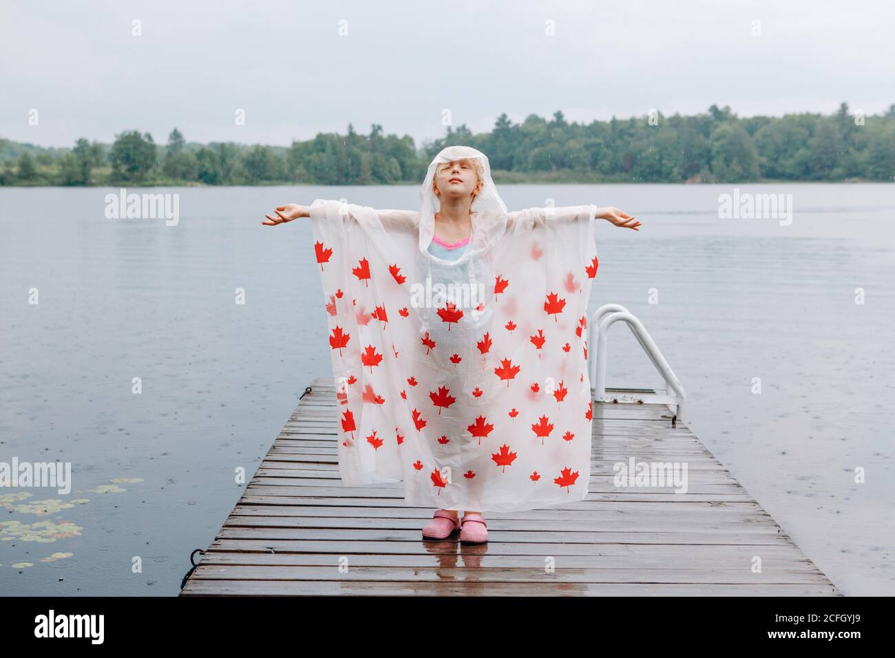 Bambina in poncho a pioggia con foglie di acero rosso in piedi su bacino lacustre in legno. Bambino che alza le braccia sotto la pioggia all'aperto. Connessione con la natura. Libertà Foto Stock