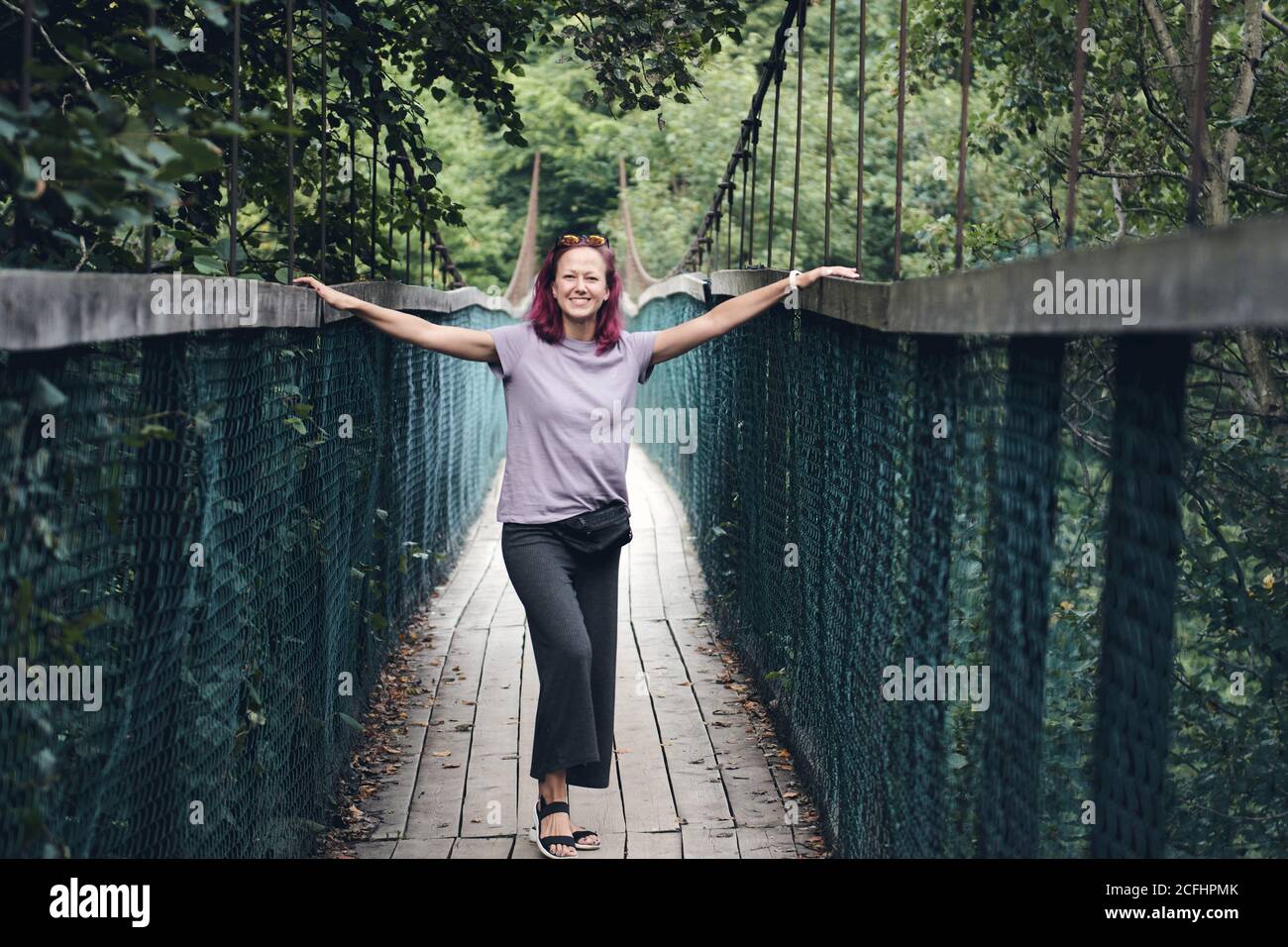 Giovane donna caucasica con capelli rosa sul ponte sospeso in acciaio lungo. Vecchio piccolo ponte di legno sul fiume solo per pedoni. Fiume di montagna Foto Stock