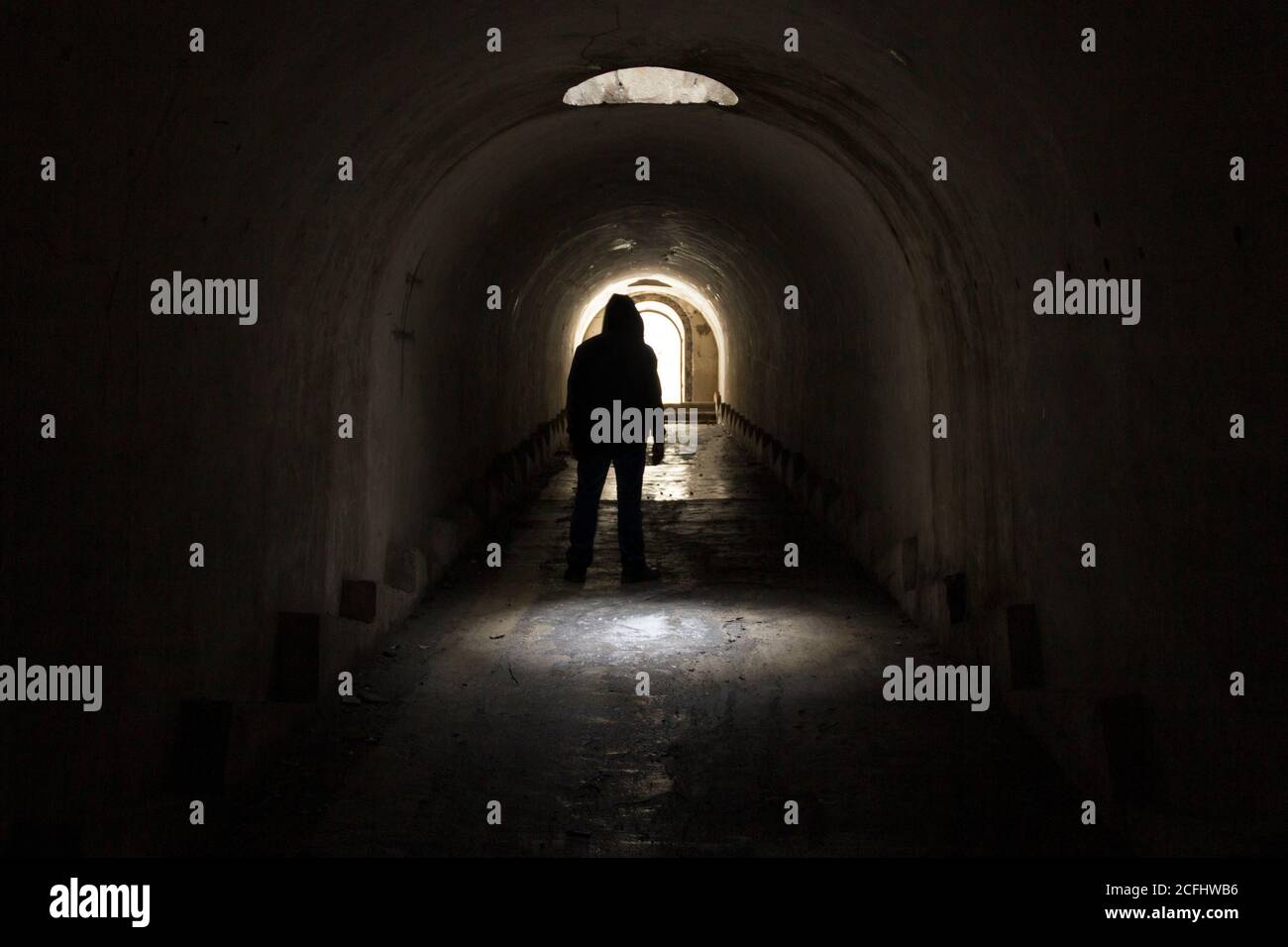 Un uomo in abiti scuri con un cappuccio che va all'uscita della luce da un tunnel sotterraneo buio e cupo, illuminato da luce solare da un portello nel soffitto Foto Stock
