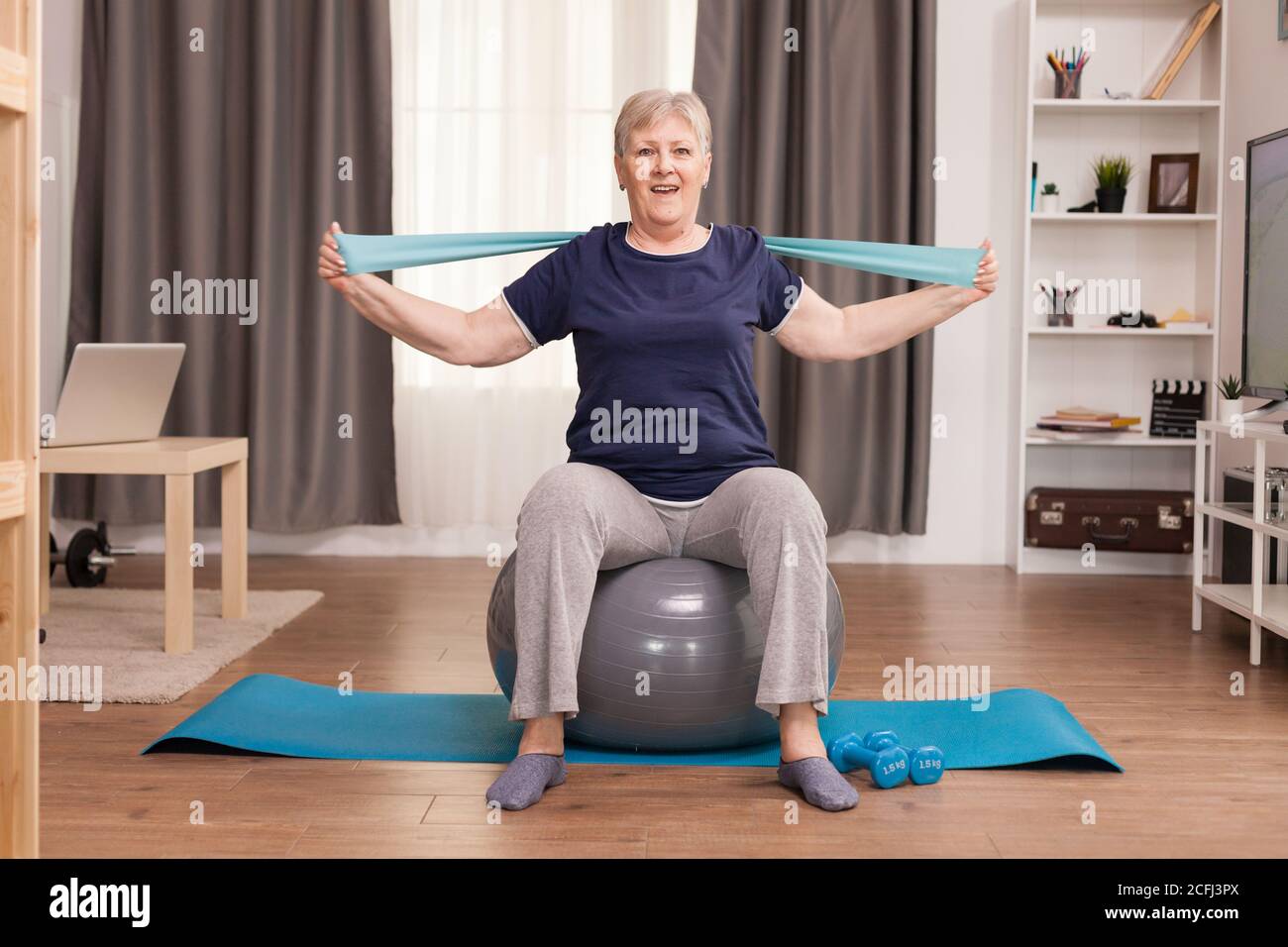 Donna anziana che si gode l'esercizio nel suo accogliente appartamento. Anziano pensionato online internet esercizio di formazione a casa attività sportiva con manubri, banda di resistenza, palla svizzera all'età di anziano pensionamento Foto Stock
