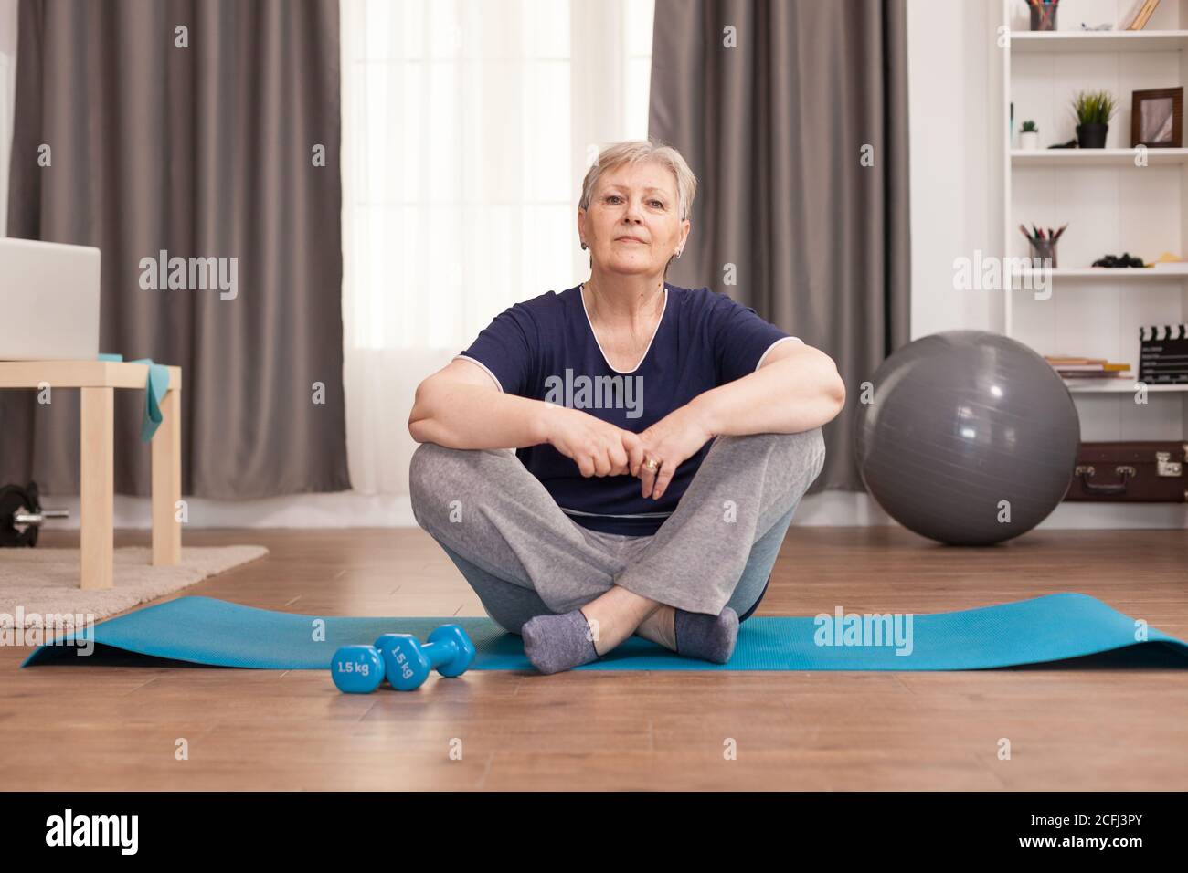 Ritratto di una donna anziana seduta sul tappetino yoga. Anziano pensionato online internet esercizio di formazione a casa attività sportiva con manubri, banda di resistenza, palla svizzera all'età di anziano pensionamento Foto Stock