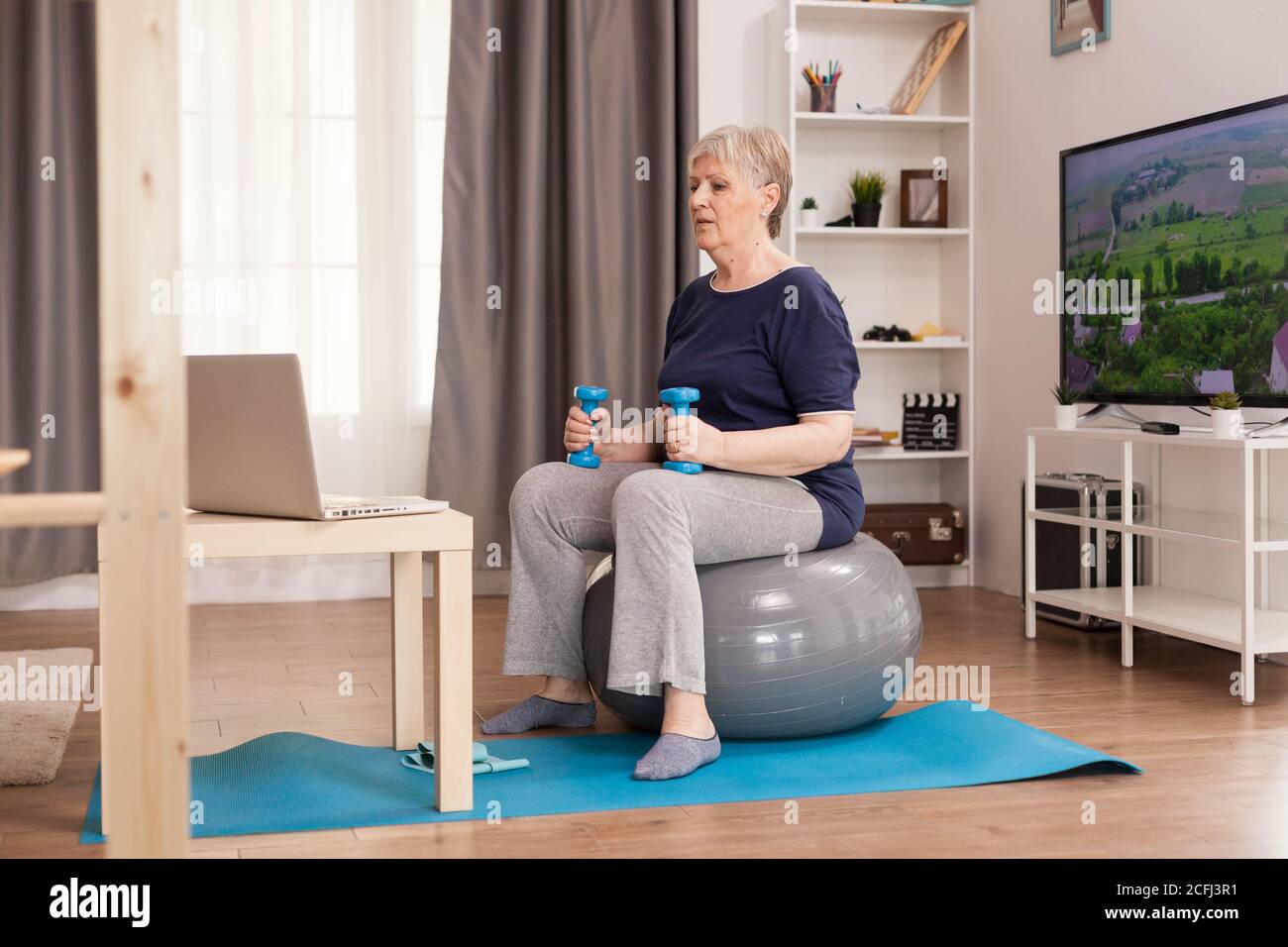 Vecchia casalinga che guarda la lezione in linea di idoneità a casa. Anziano pensionato online internet esercizio di formazione a casa attività sportiva con manubri, banda di resistenza, palla svizzera all'età di anziano pensionamento Foto Stock