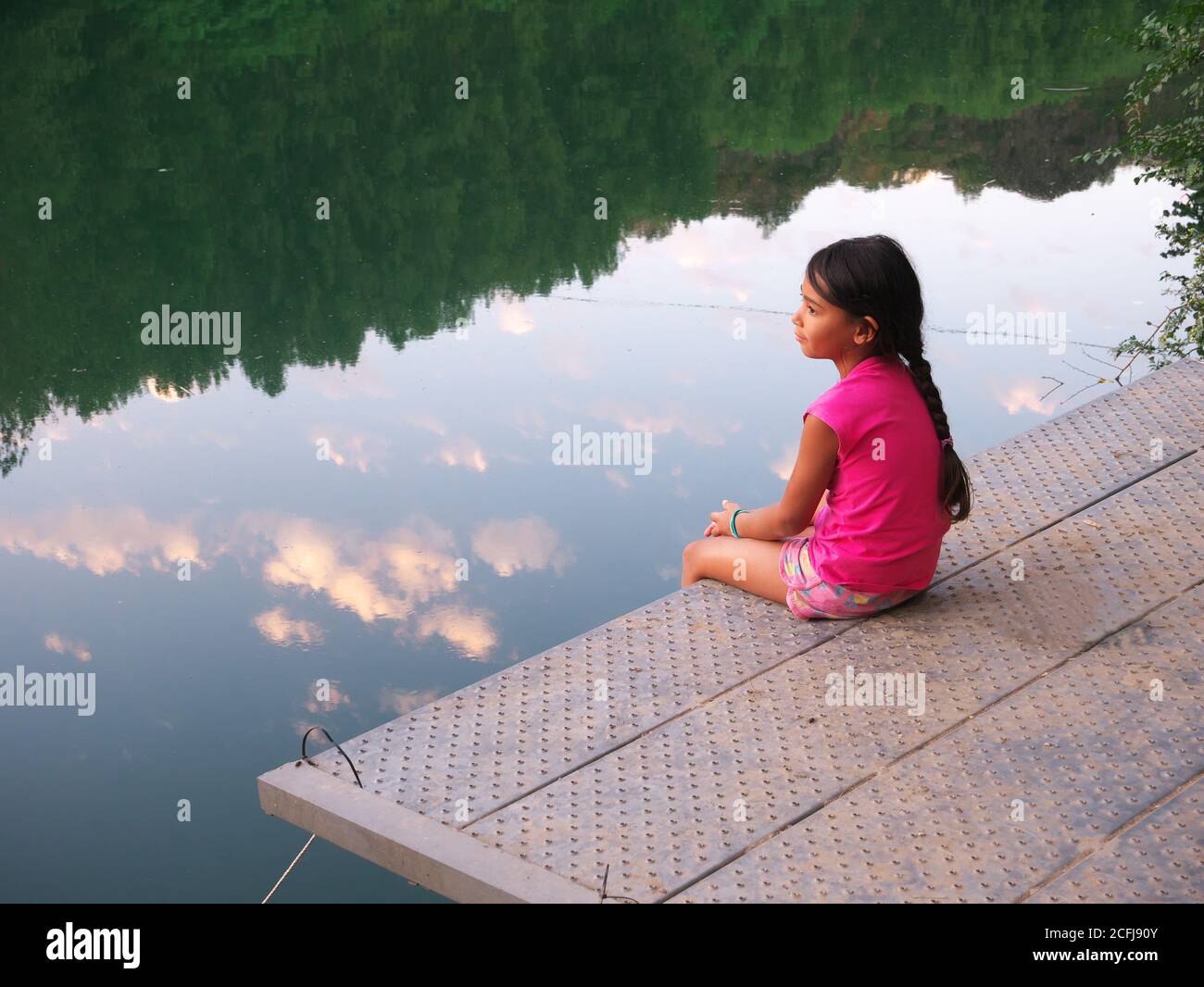 Bambina penosa vista in profilo seduta su un pontile vicino al fiume, contemplando la natura Foto Stock