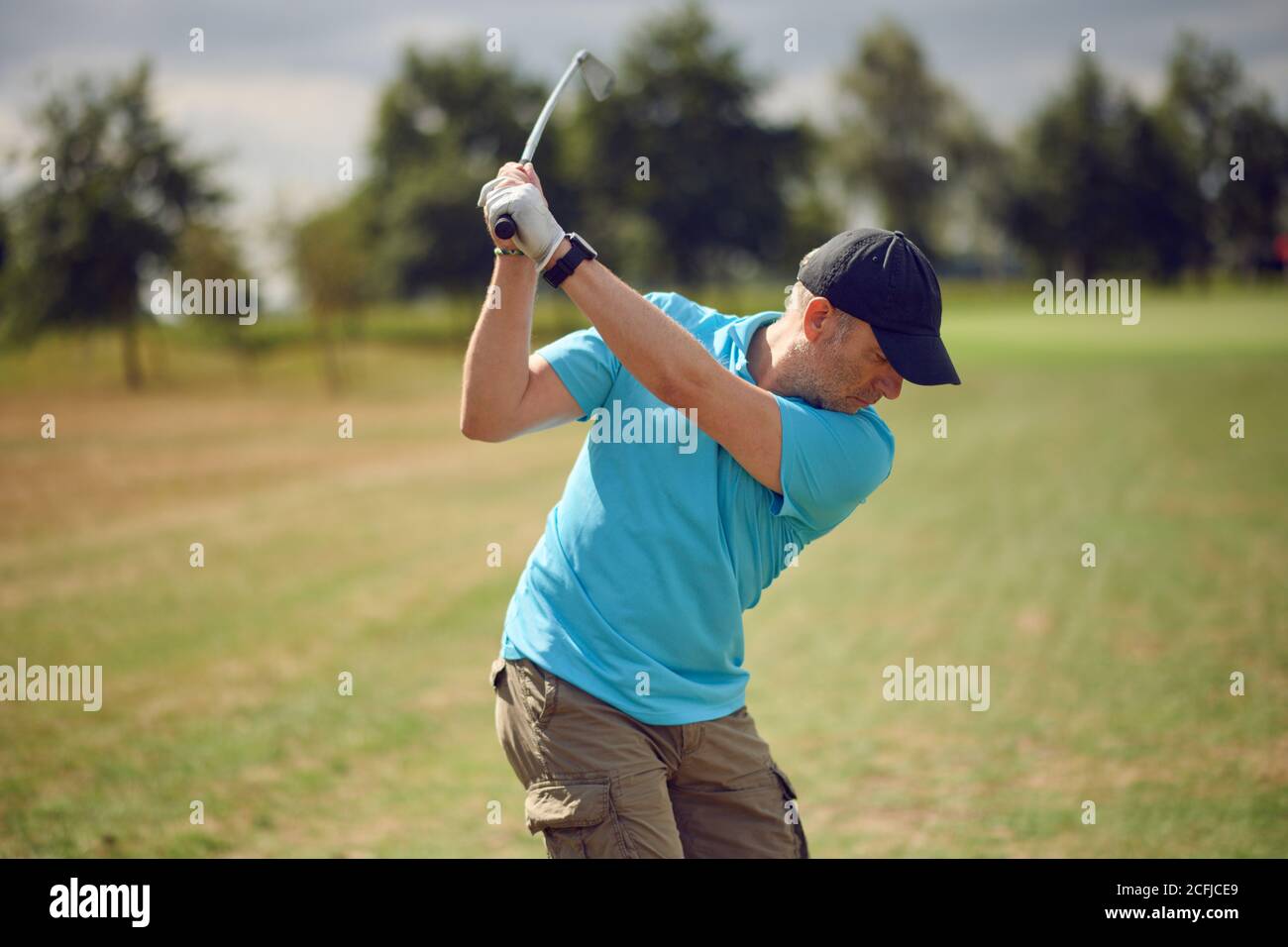 Uomo che gioca a golf oscillando alla palla come gioca il suo scatto utilizzando un driver visto da dietro guardando verso il basso il fairway in un sano stile di vita attivo co Foto Stock