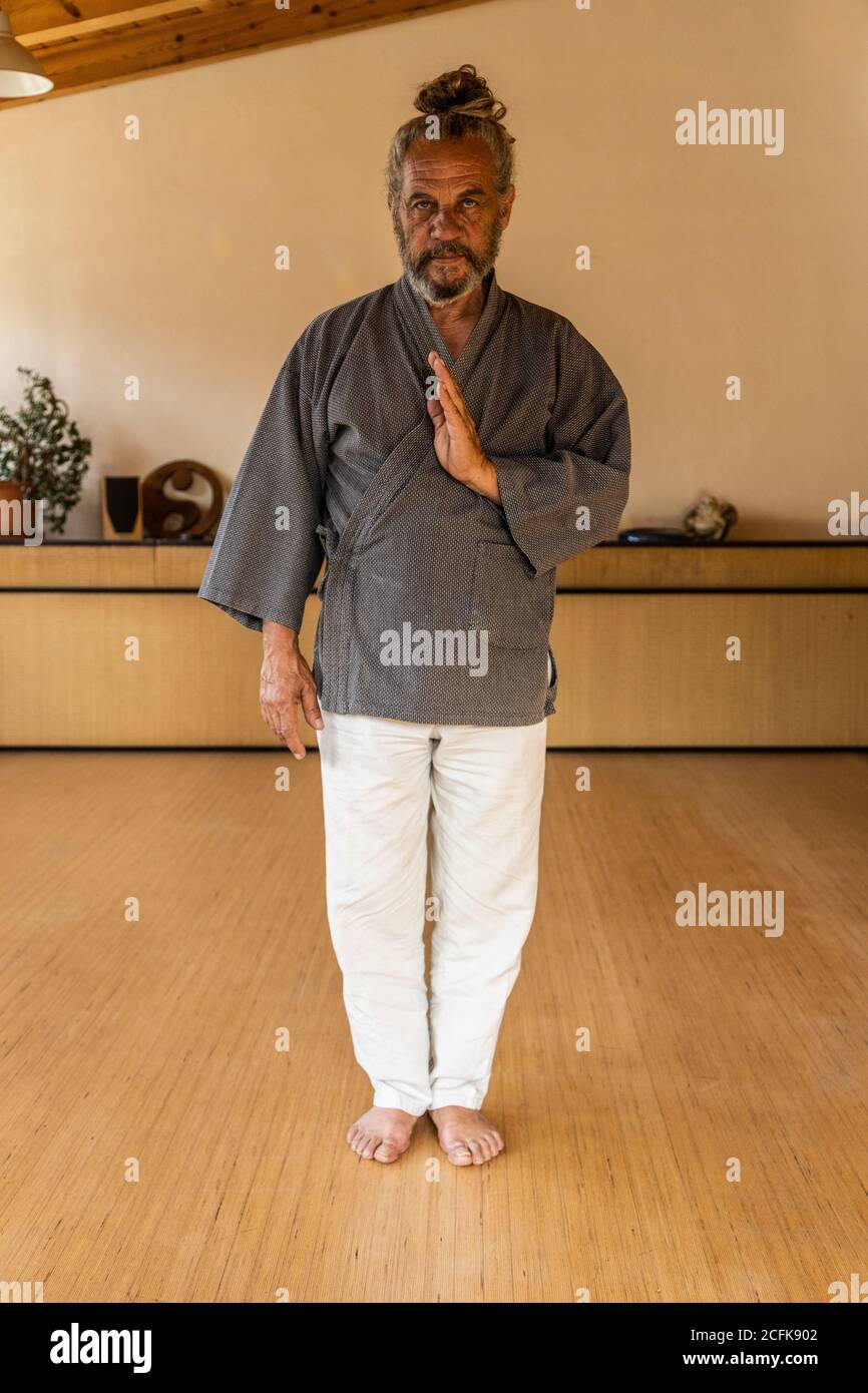 Anziano portatore di chi kung maschio etnico in abbigliamento tradizionale guardando la fotocamera mentre si è in piedi in uno studio luminoso Foto Stock
