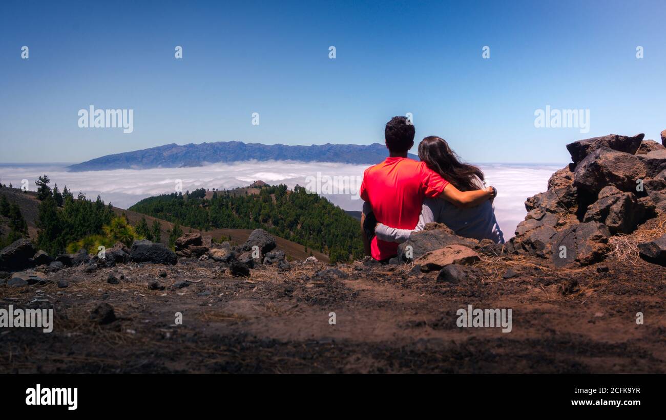 Vista posteriore della coppia di viaggio tenero seduta su una collina e. abbracciare mentre si ammira il paesaggio spettacolare sulle nuvole su terreni montuosi Foto Stock