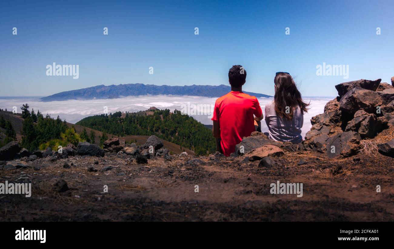 Vista posteriore della coppia di viaggio tenero seduta su una collina e. abbracciare mentre si ammira il paesaggio spettacolare sulle nuvole su terreni montuosi Foto Stock