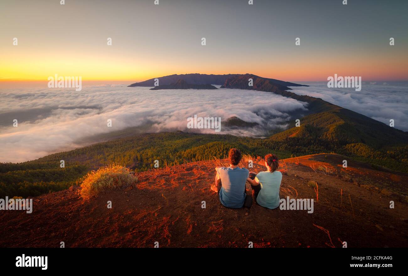 Vista posteriore della coppia di viaggio tenero seduto in collina mentre ammira lo spettacolare paesaggio sulle nuvole su terreni montuosi Foto Stock