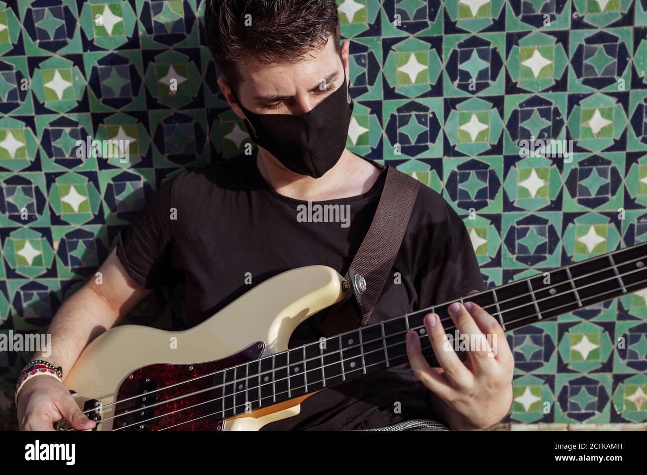 Corpo pieno di giovane uomo in maschera protettiva nera per prevenzione del coronavirus seduto sui gradini vicino alla parete e suonando il basso chitarra in strada Foto Stock