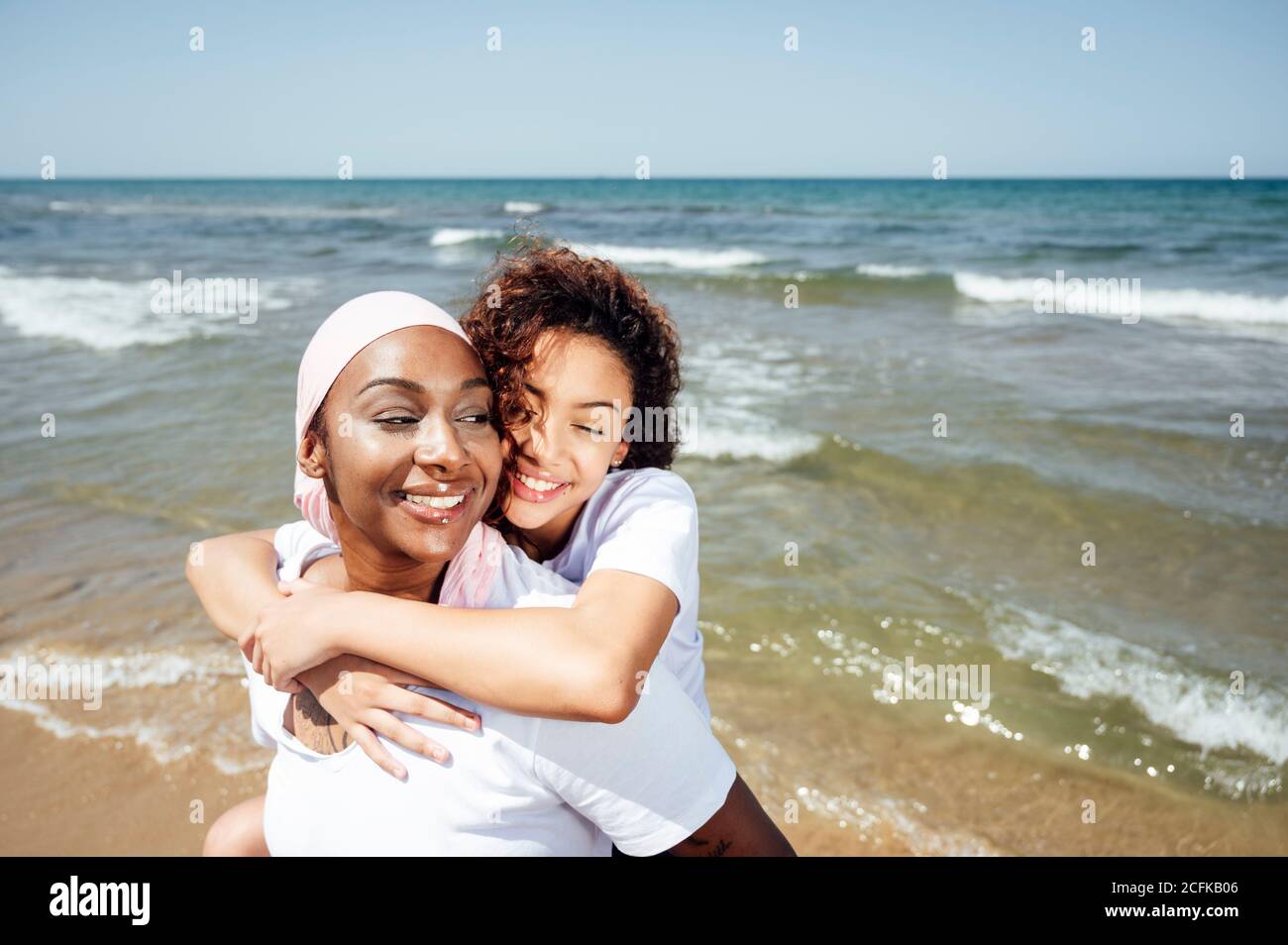 Felice madre nera piggybacking giovane figlia mentre si divertono con occhi chiusi al mare in estate Foto Stock