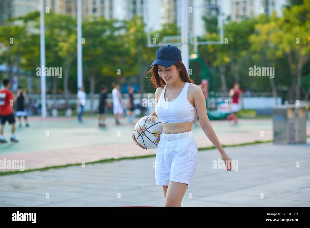 Spensierata donna asiatica in sportswear che cammina con il basket lungo gli sport terreno in estate e guardando verso il basso Foto Stock