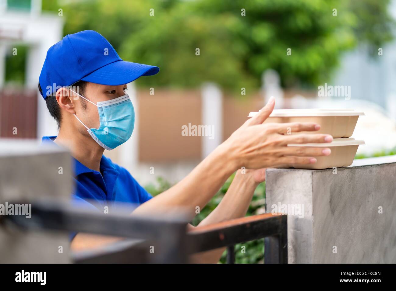 Asian consegnare l'uomo con maschera facciale in camicia blu maneggiando scatole di cibo e messo sul posto recinzione della casa del cliente come consegna di cibo senza contatto. Questo c Foto Stock