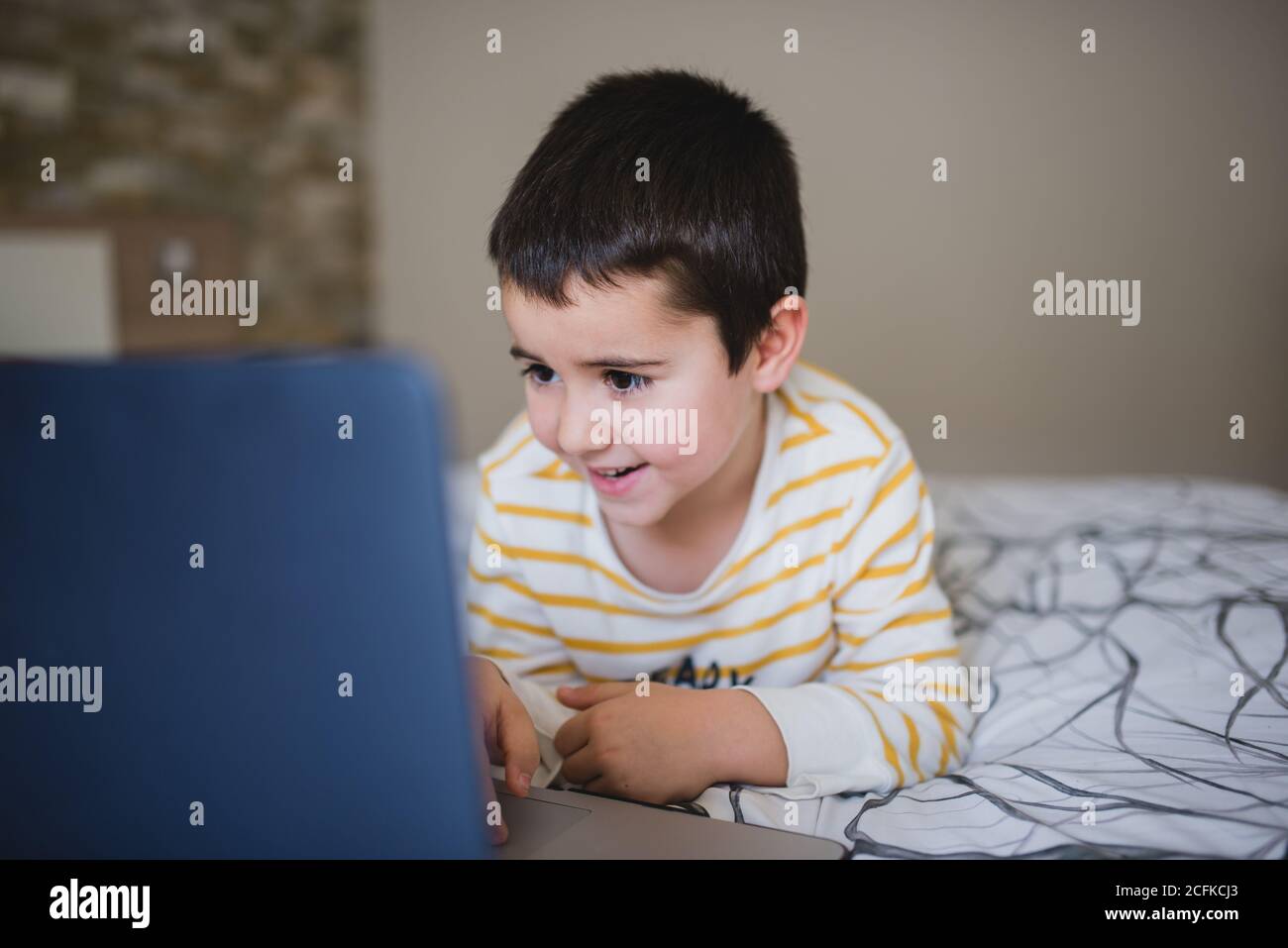 Piccolo ragazzo concentrato in abbigliamento casual che naviga il laptop durante la spesa tempo libero in camera da letto a casa Foto Stock