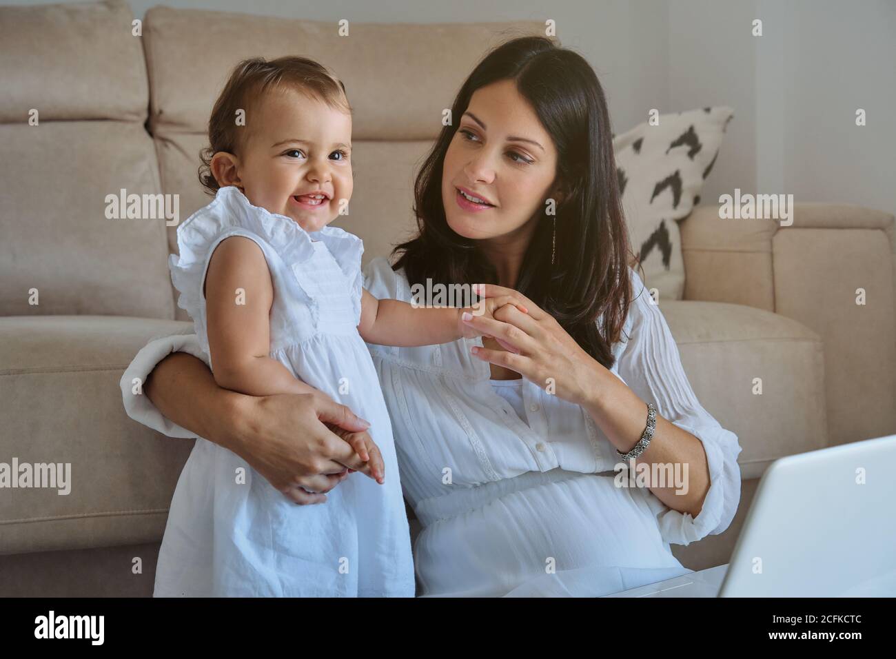 Bambina in un vestito bianco sorridente in piedi mentre sua madre si siede sul pavimento con un aspetto tenero accanto al divano nella casa Foto Stock