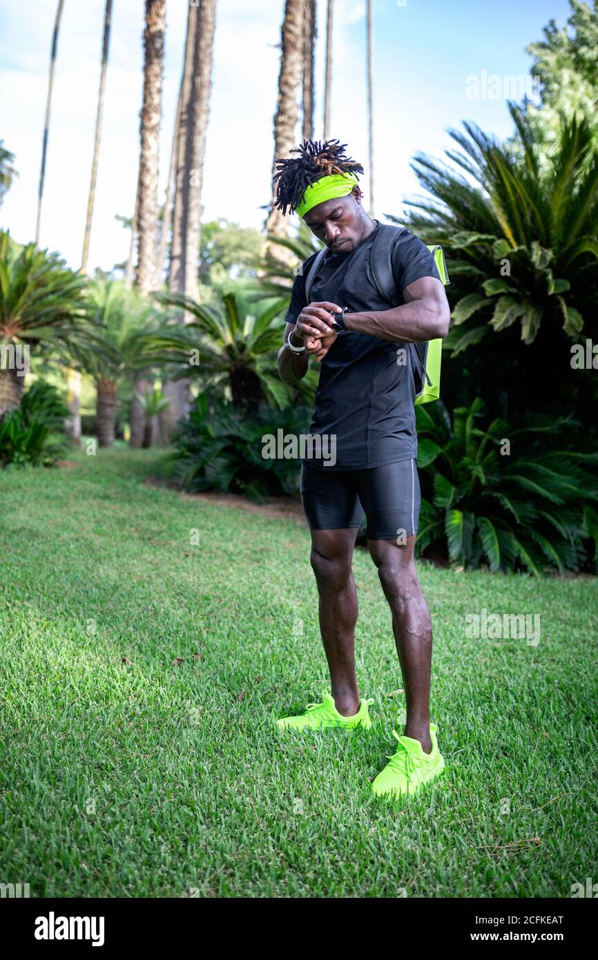 Giovane ed elegante corridore africano-americano con cuffie sul collo controllare i risultati sportivi sul tracker fitness durante l'allenamento in verde parco tropicale Foto Stock