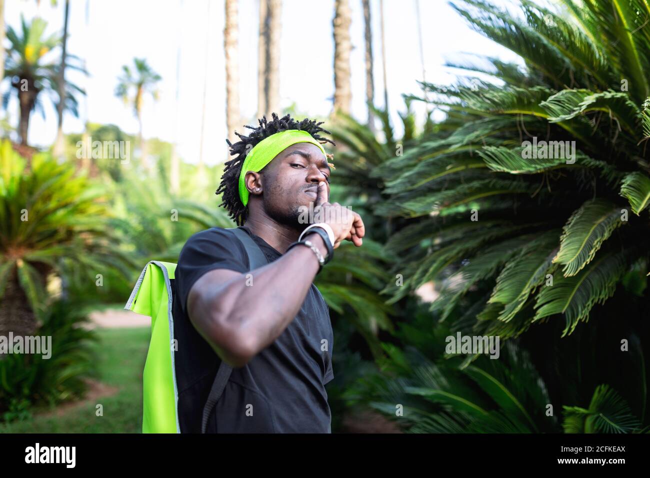 Giovane atleta africano-americano maschile in abbigliamento sportivo e per la fabbricazione di fasce shh gesto mentre si è in piedi contro piante tropicali verdi sulla strada Foto Stock