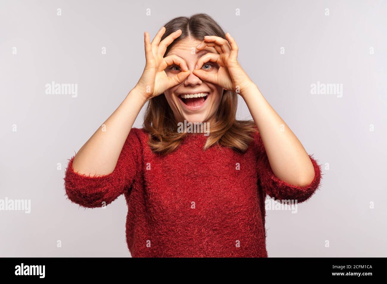 Ritratto di ragazza felice spensierata in maglione shaggy guardando attraverso i buchi delle dita, binocolo gesto della mano, facendo occhiali e guardando con eccitatori Foto Stock