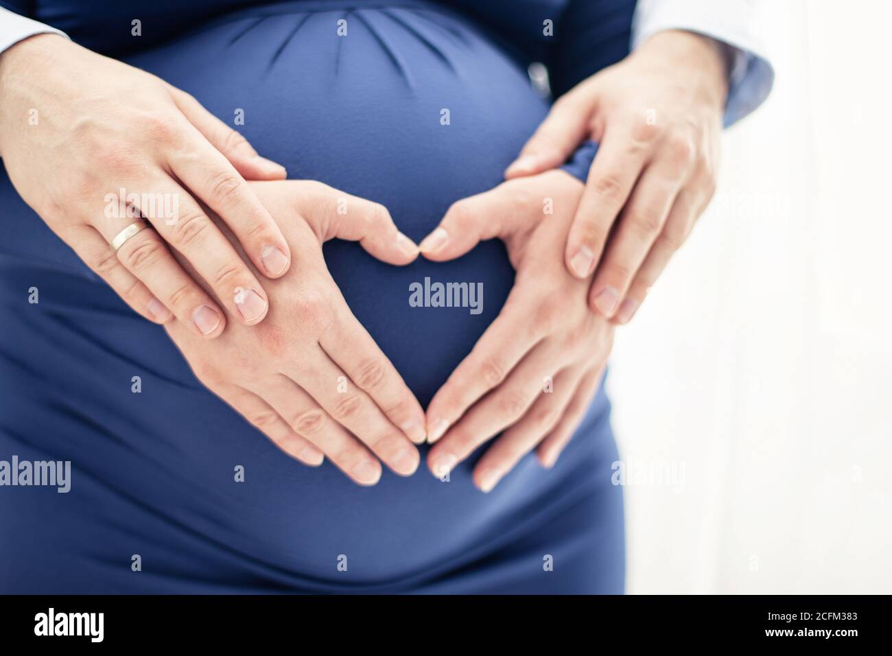 Le mani dell'uomo toccano le mani della donna incinta a forma di cuore sul suo bump del bambino. Foto luminosa su sfondo bianco Foto Stock