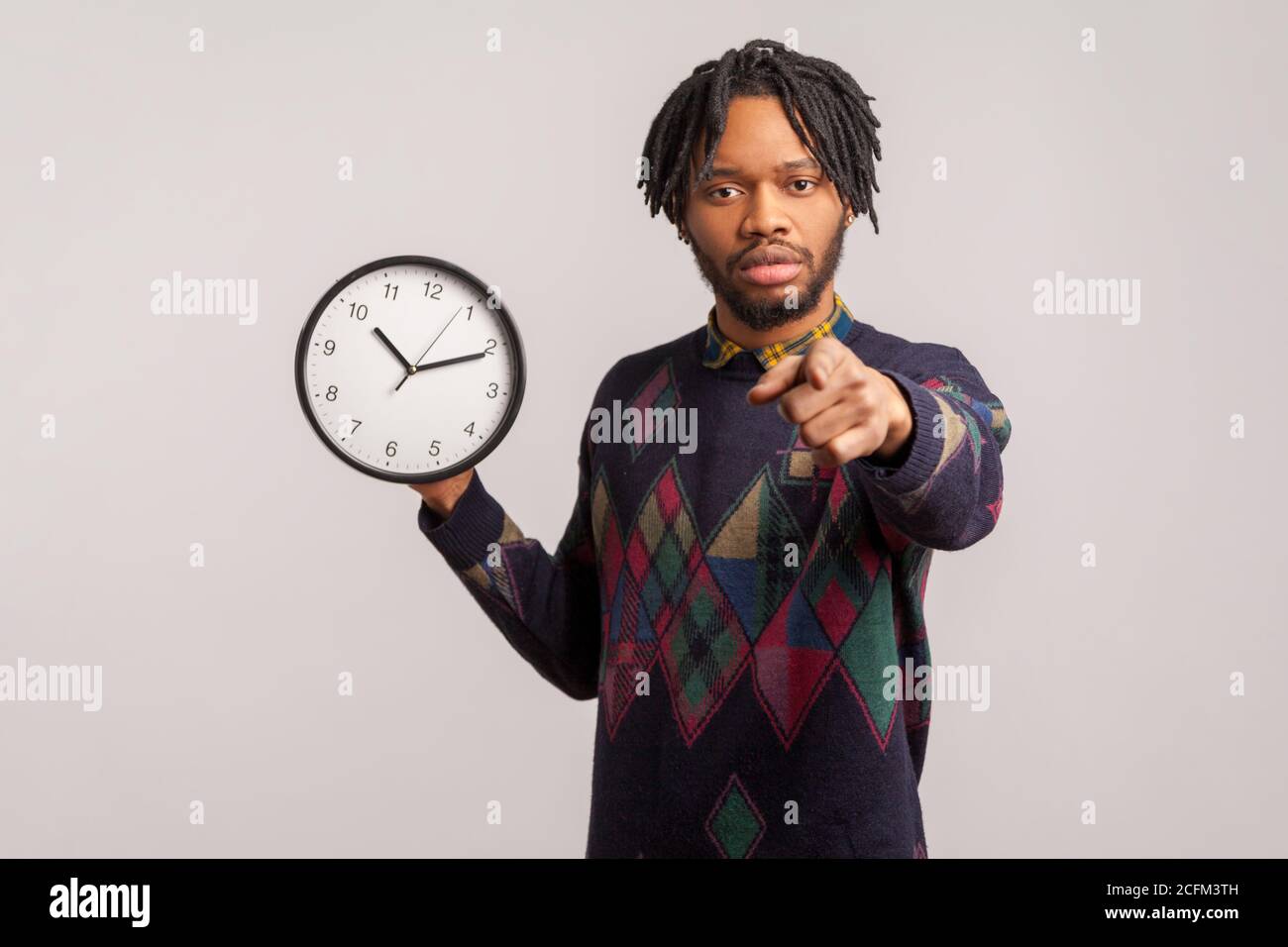 Uomo africano serio bossy con i Dreadlock che tengono l'orologio da parete in mano che punta il dito alla macchina fotografica con lo sguardo concentrato, il tempo all'azione, la motivazione. IND Foto Stock