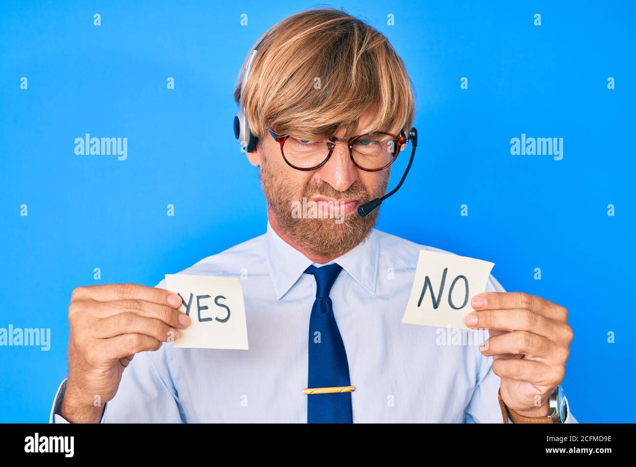 Giovane biondo uomo che indossa l'auricolare operatore che tiene sì e nessun promemoria scettico e nervoso, rondante sconvolto a causa del problema. Persona negativa. Foto Stock
