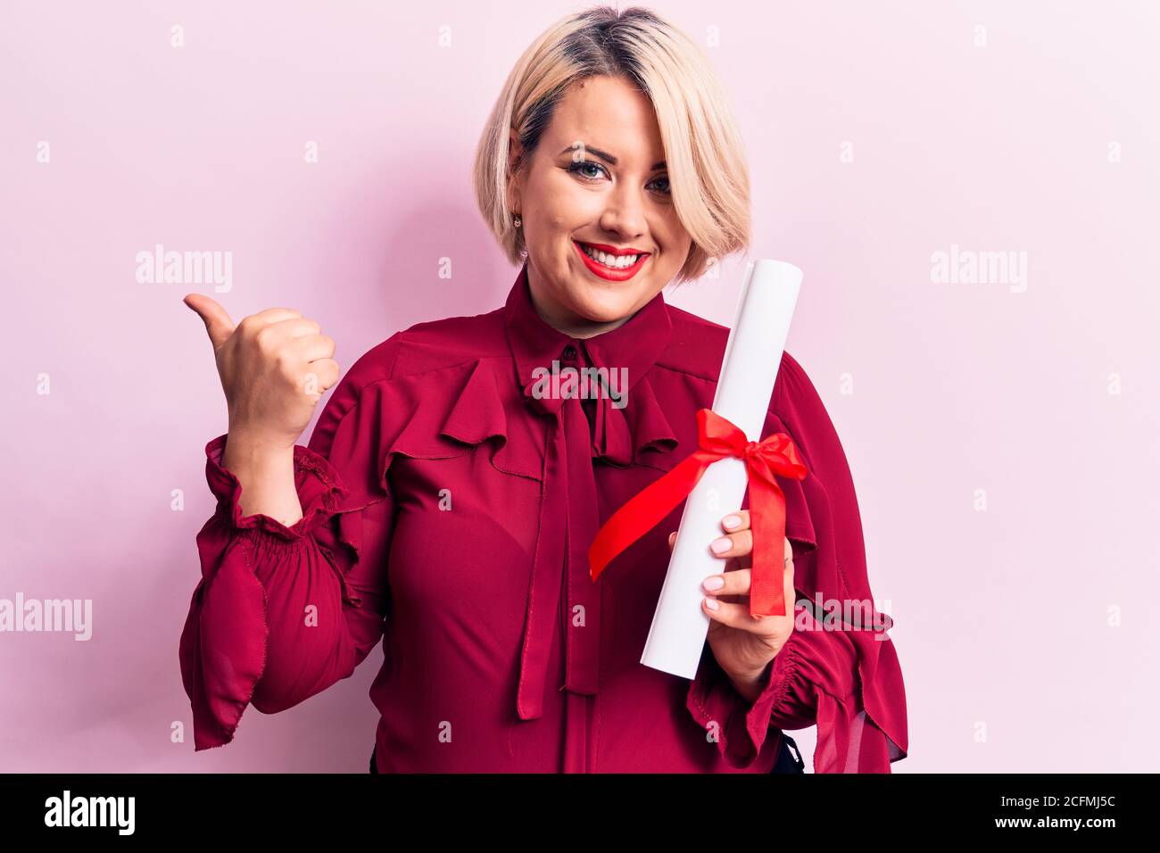 Bella bionda più la taglia donna che tiene il diploma laureato sopra sfondo rosa che punta il pollice verso il lato sorridendo felice a bocca aperta Foto Stock