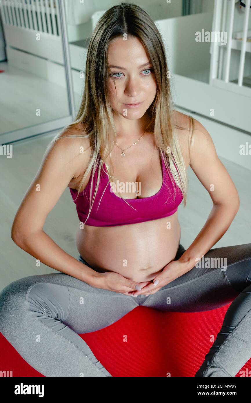 Donna incinta che si alleva yoga, carezzandogli la pancia seduta sul tappeto rosso yoga allenamento meditazione yoga. Foto Stock